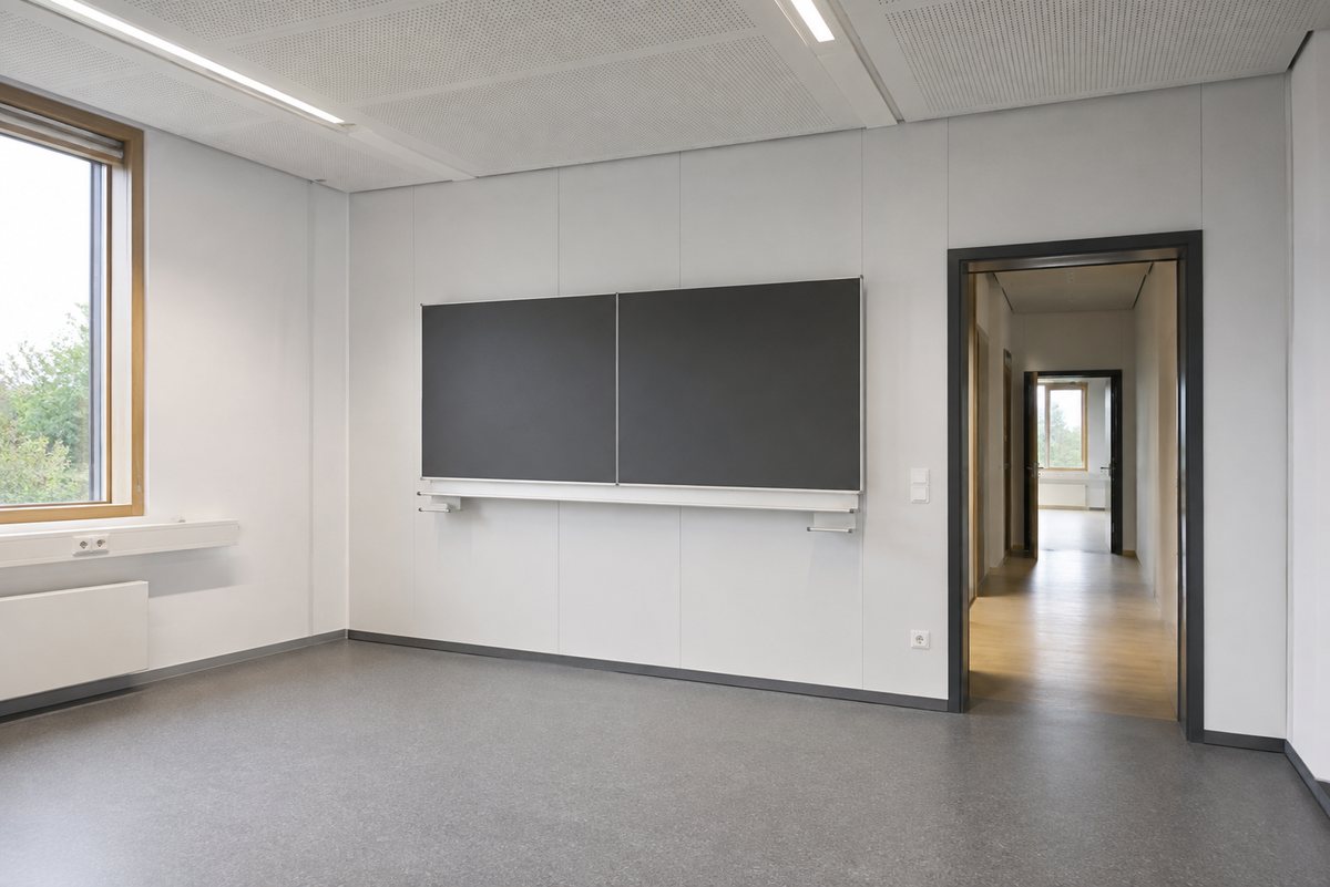 Leeres Klassenzimmer mit weißen Wandplatten, grauem Boden, Akustikdecke und zweiteiliger dunkler Tafel; rechts der Blick durch die offene Tür in den Flur
