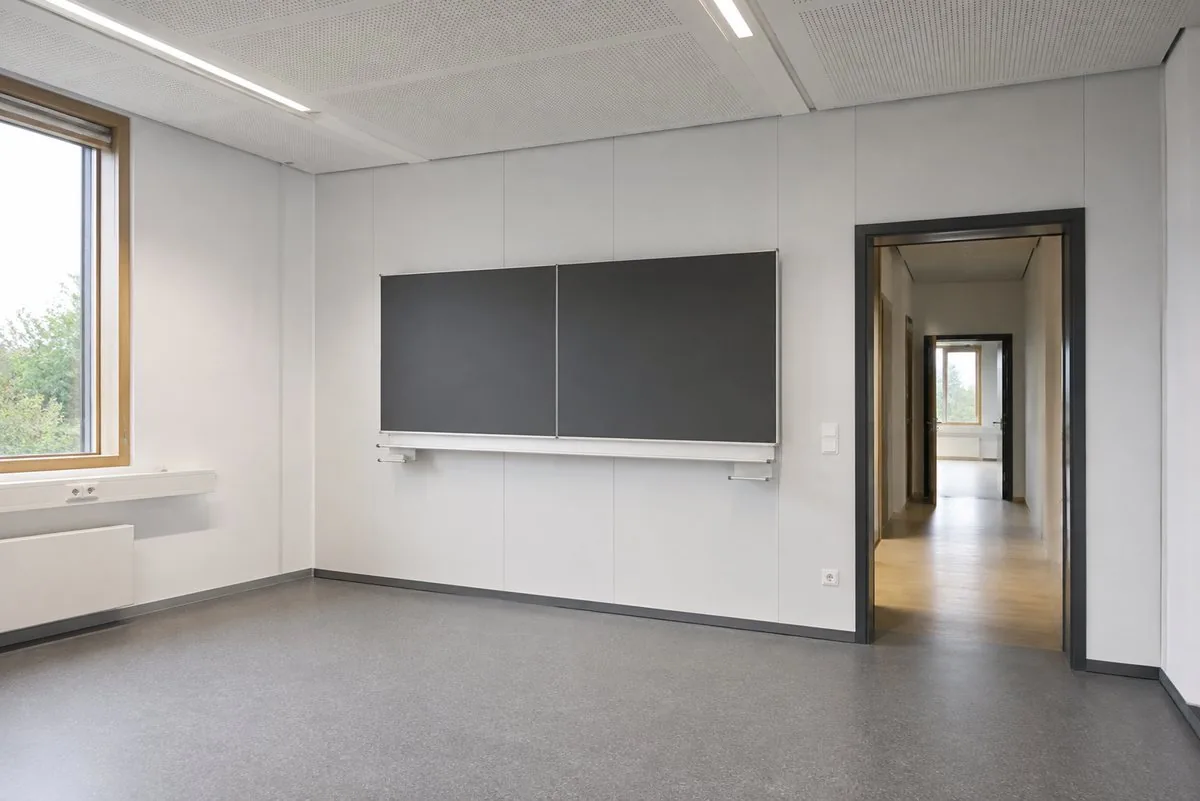 Leeres Klassenzimmer mit weißen Wandplatten, grauem Boden, Akustikdecke und zweiteiliger dunkler Tafel; rechts der Blick durch die offene Tür in den Flur
