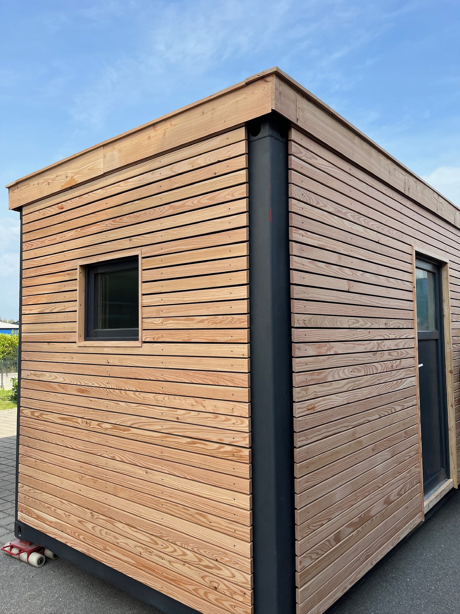 Gartenhaus 15 m² von Contain-us – Außenansicht mit Holzfassade und kleinem Fenster, das Modul steht auf einer gepflasterten Fläche