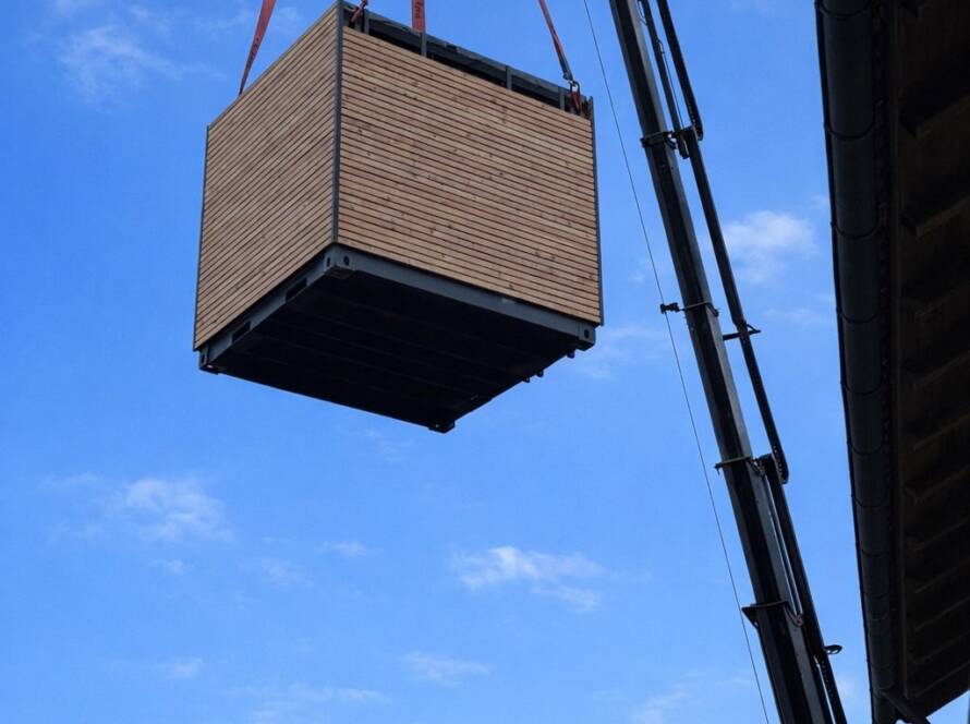Ein Autokran hebt ein fertiges Contain-us Modul mit Holzfassade durch die Luft, darunter Wohnhäuser