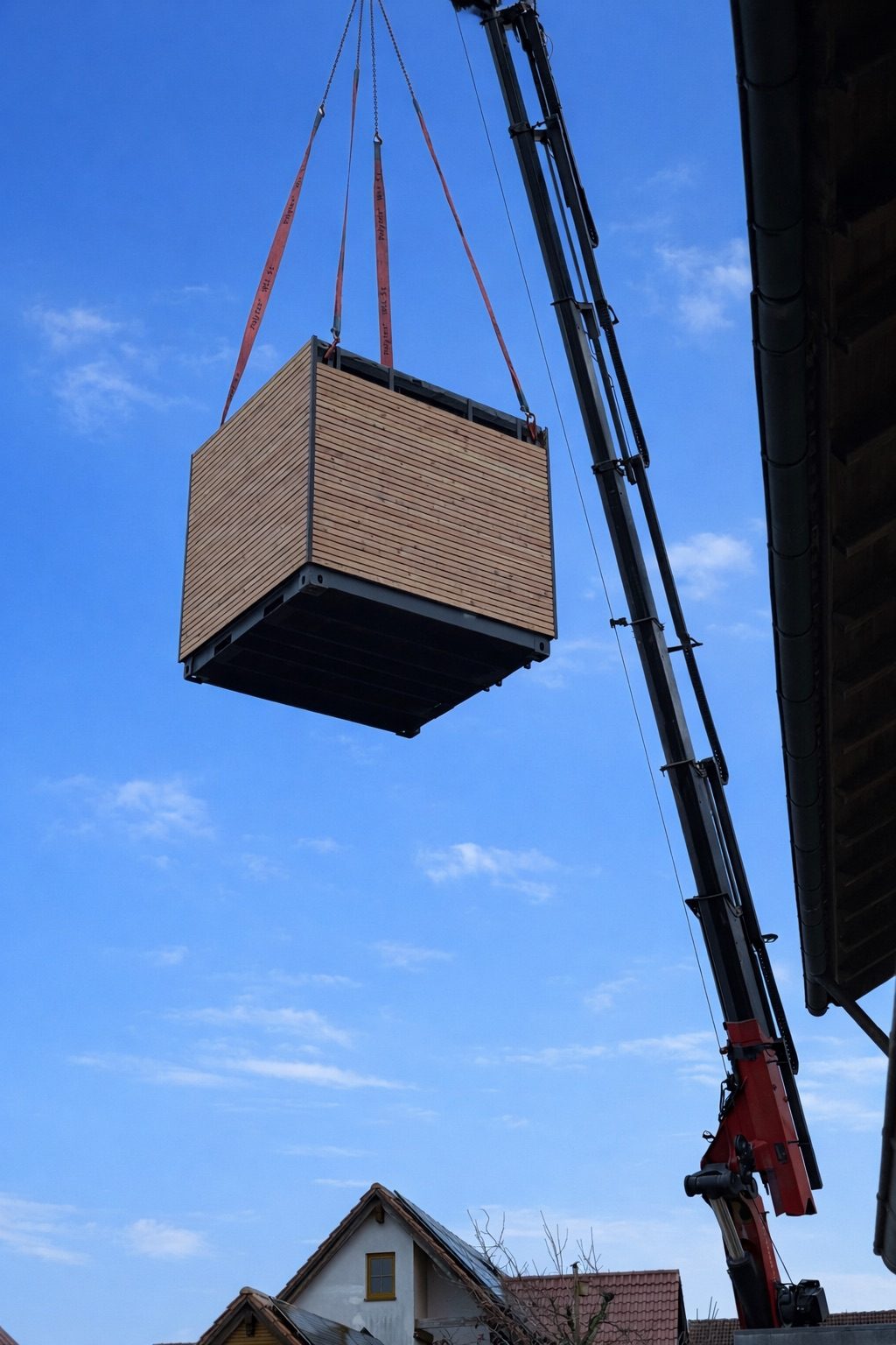 Ein Autokran hebt ein fertiges Contain-us Modul mit Holzfassade durch die Luft, darunter Wohnhäuser