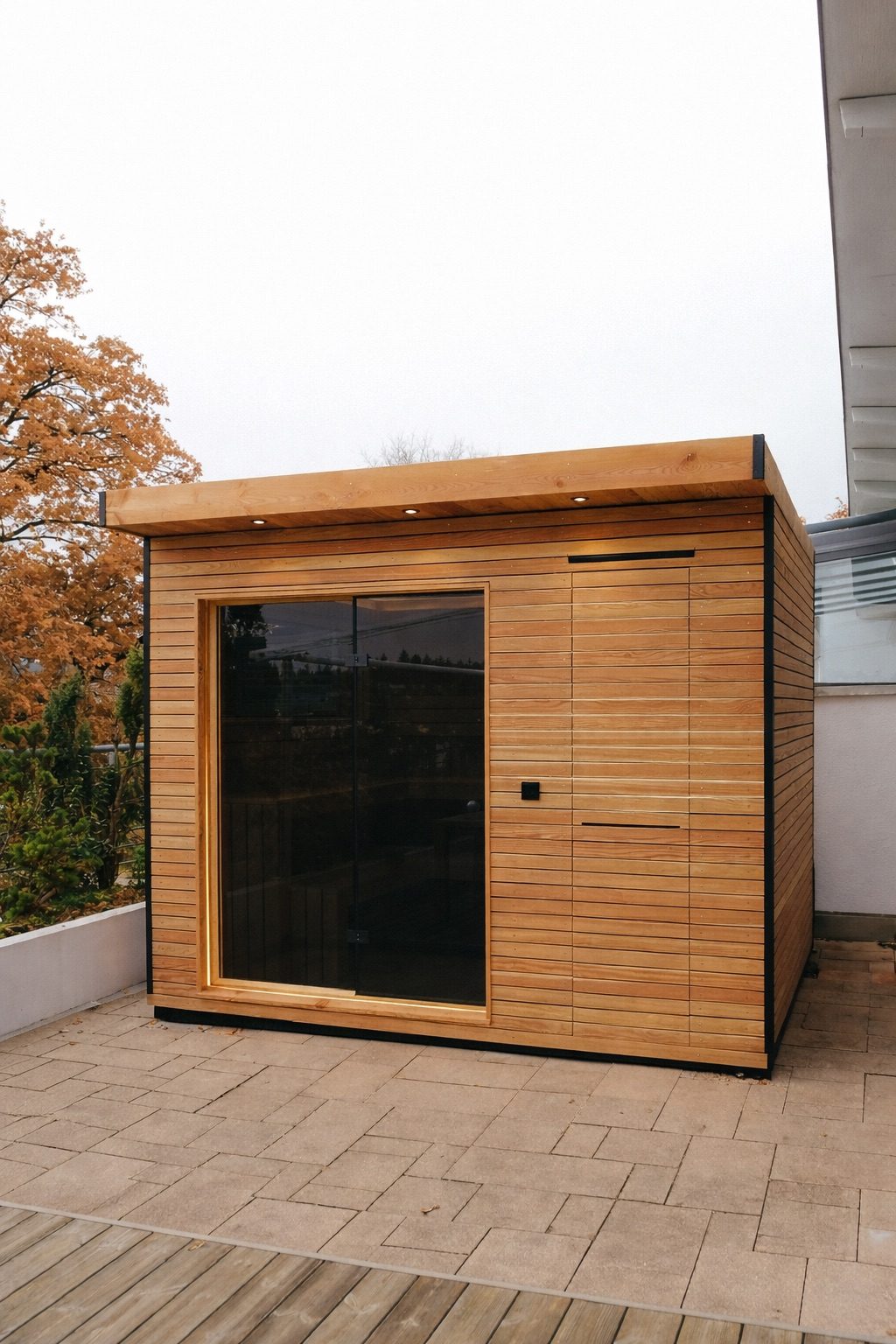 Sauna 7 m² von Contain-us – Außenansicht der Gartensauna auf einer gepflasterten Fläche, daneben ein Fenster zum Garten