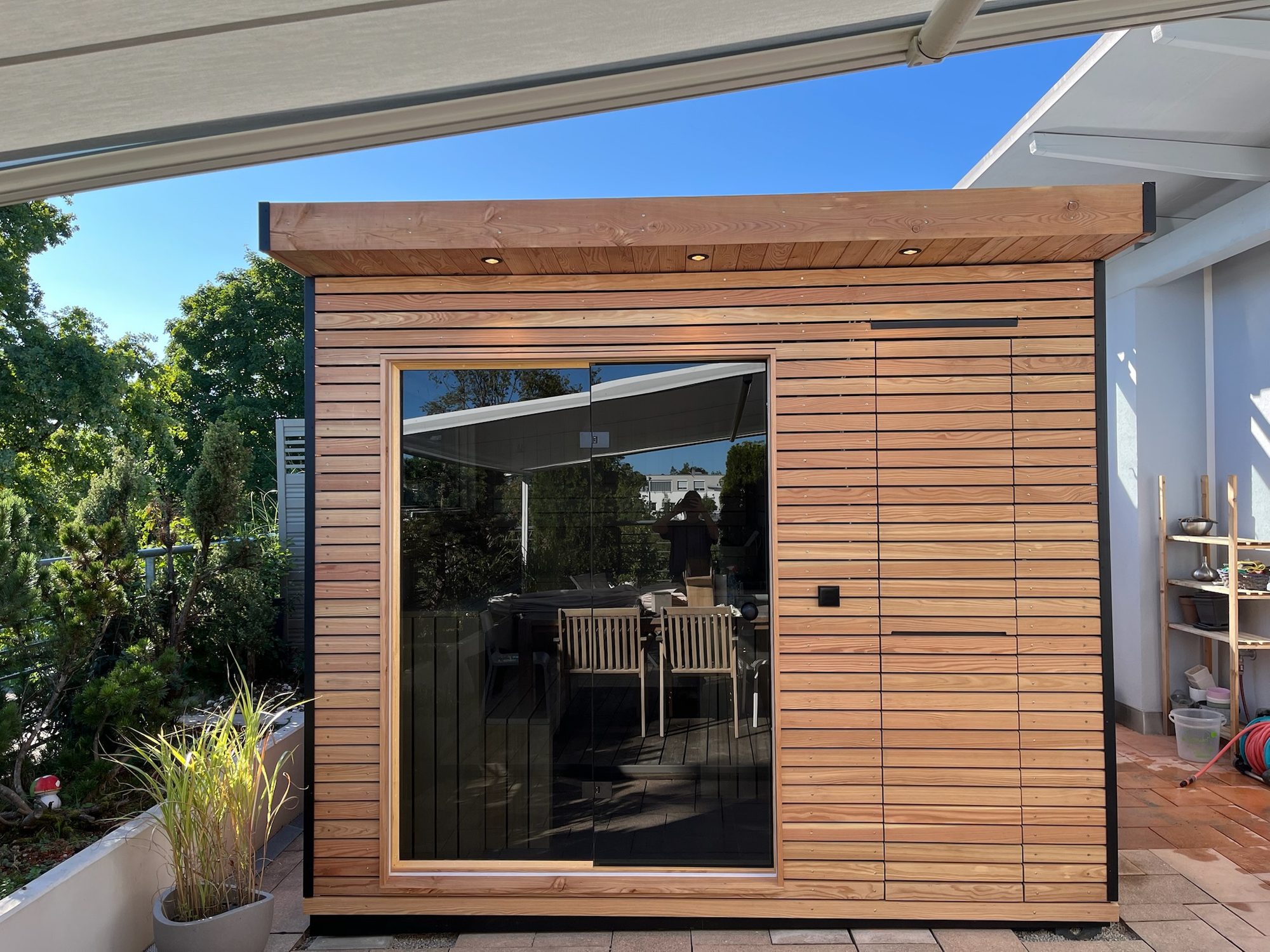 Sauna 7 m² von Contain-us – Außenansicht unter einem Vordach, Schiebetür und Holzlamellen