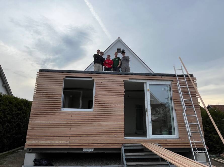 Modul auf einem Streifenfundament mit Kiesbett, das Montageteam steht auf dem Dach, die Fassade ist teilweise verlattet