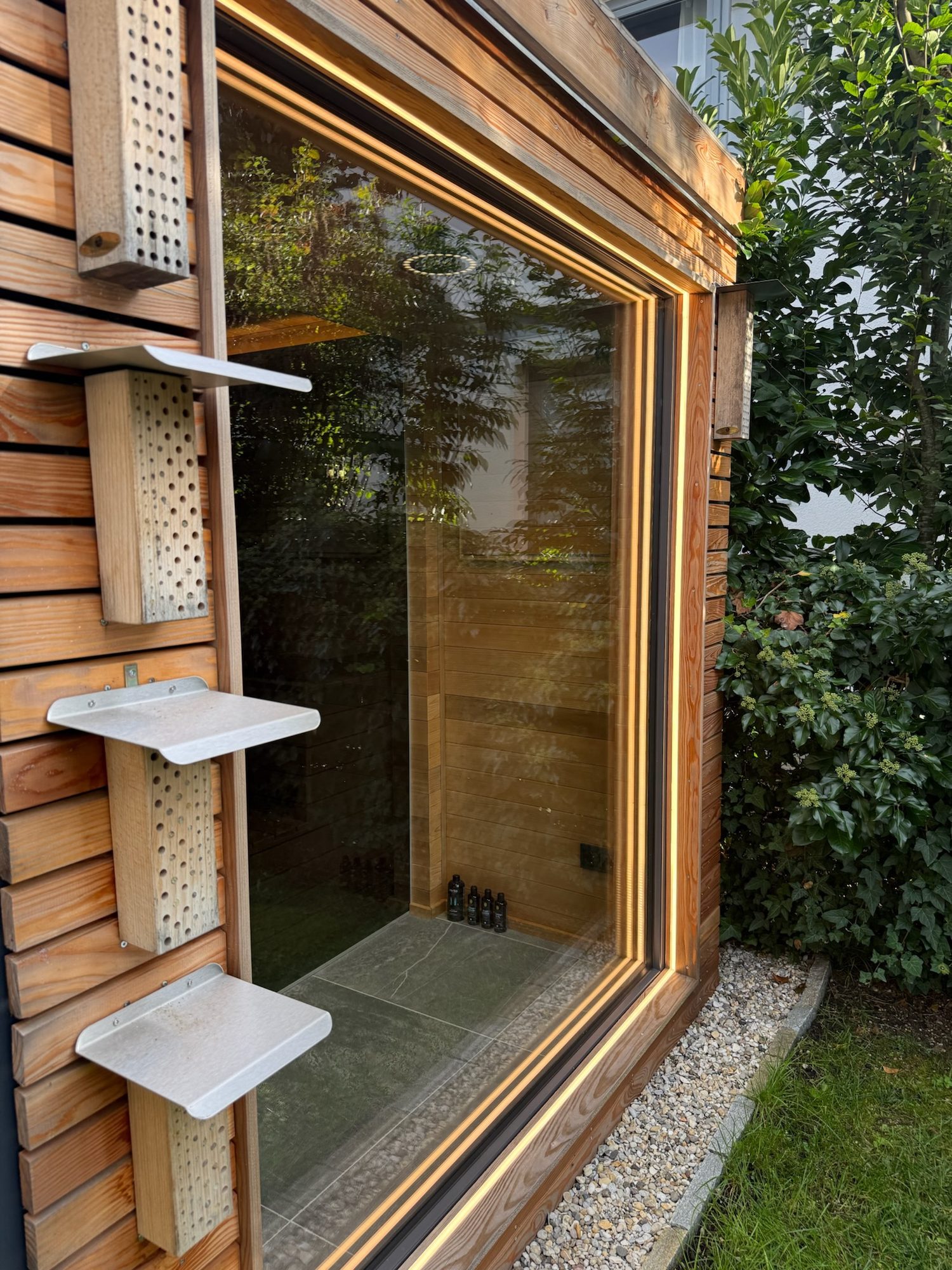 Sauna 7 m² von Contain-us – Insektenhotel mit Ablagen an der Holzfassade neben dem großen Fenster, dahinter der geflieste Duschbereich, am Sockel ein Kiesstreifen