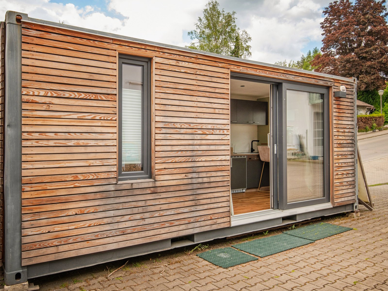 Tiny House 15 m² von Contain-us – Außenansicht mit Fensterband und geöffneter Schiebetür, davor eine gepflasterte Fläche