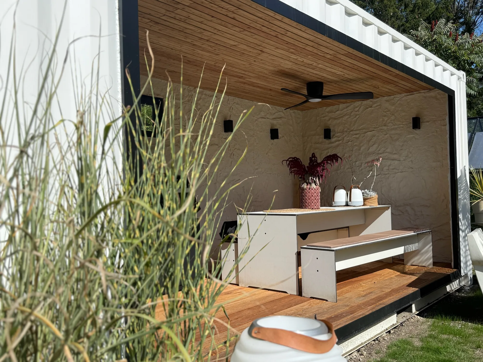 Outdoor-Küche im Modul von Contain-us – offene Front mit Lärchenholzdecke, weißem Esstisch mit Bank und Ziergras im Garten