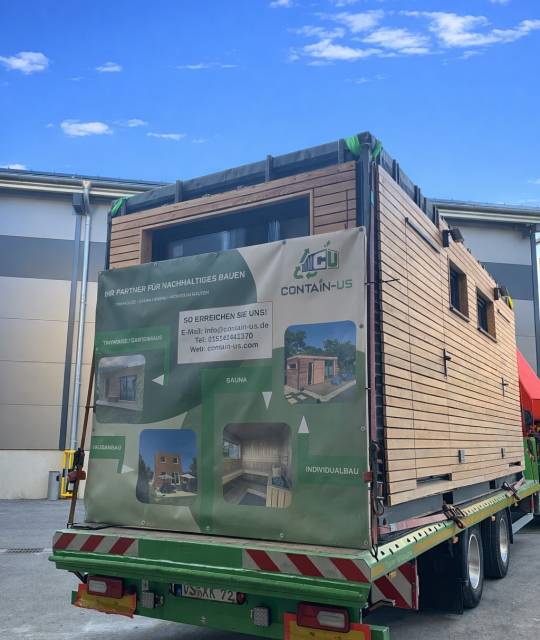 Fertiges Modul mit Lärchenholzfassade auf einem Tieflader vor der Halle in Landsberg am Lech, an der Ladefläche hängt eine Contain-us Plane