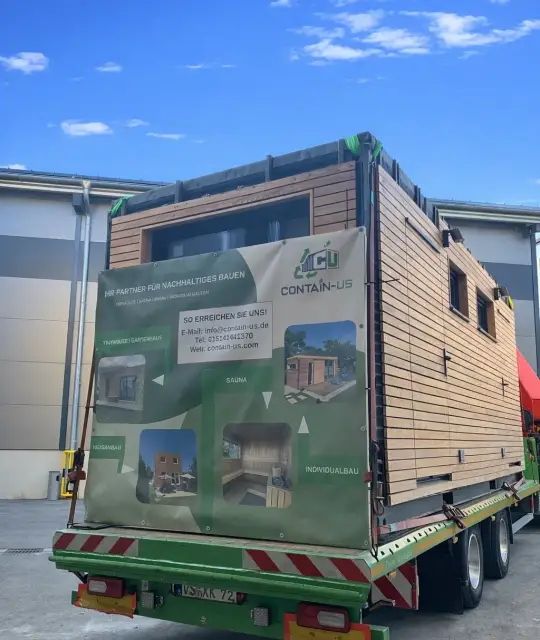 Fertiges Modul mit Lärchenholzfassade auf einem Tieflader vor der Halle in Landsberg am Lech, an der Ladefläche hängt eine Contain-us Plane